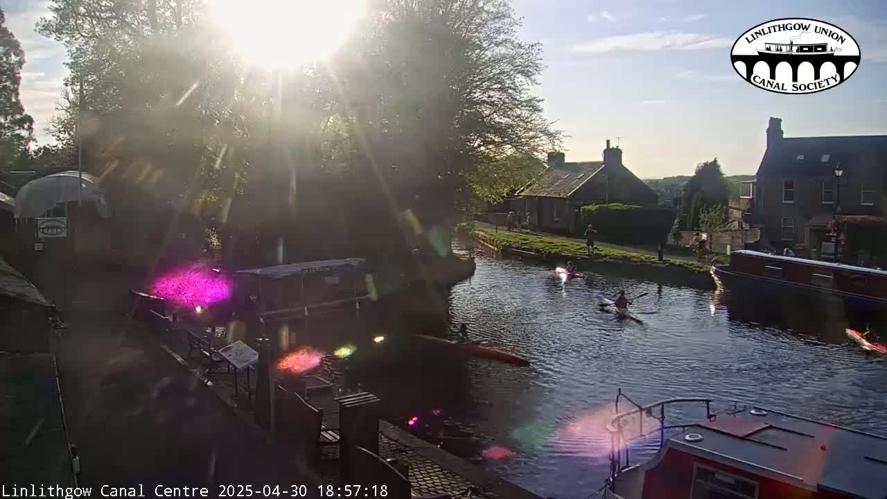 Kayakers paddle on a calm canal under a bright, sunny sky, with buildings and trees lining the waterway.