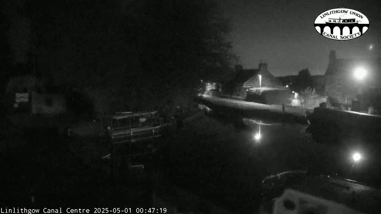 A nighttime view of a canal, showing calm water reflecting lights from nearby buildings and boats moored along the bank.