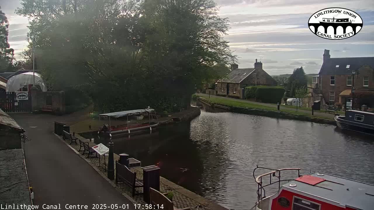 A canal scene under a partly cloudy sky shows boats moored along its banks, near stone buildings and lush green trees.