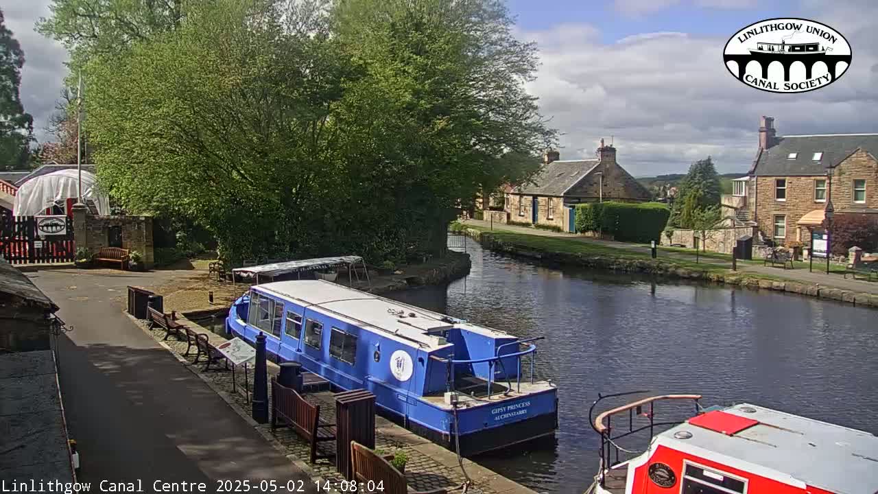 A blue narrowboat is docked at a canal side, with stone buildings and lush greenery visible on a partly cloudy day.