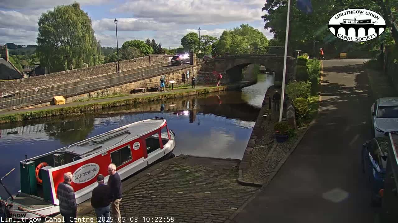 A red canal boat is moored near a stone bridge and towpath, with several people standing nearby under a sunny sky.