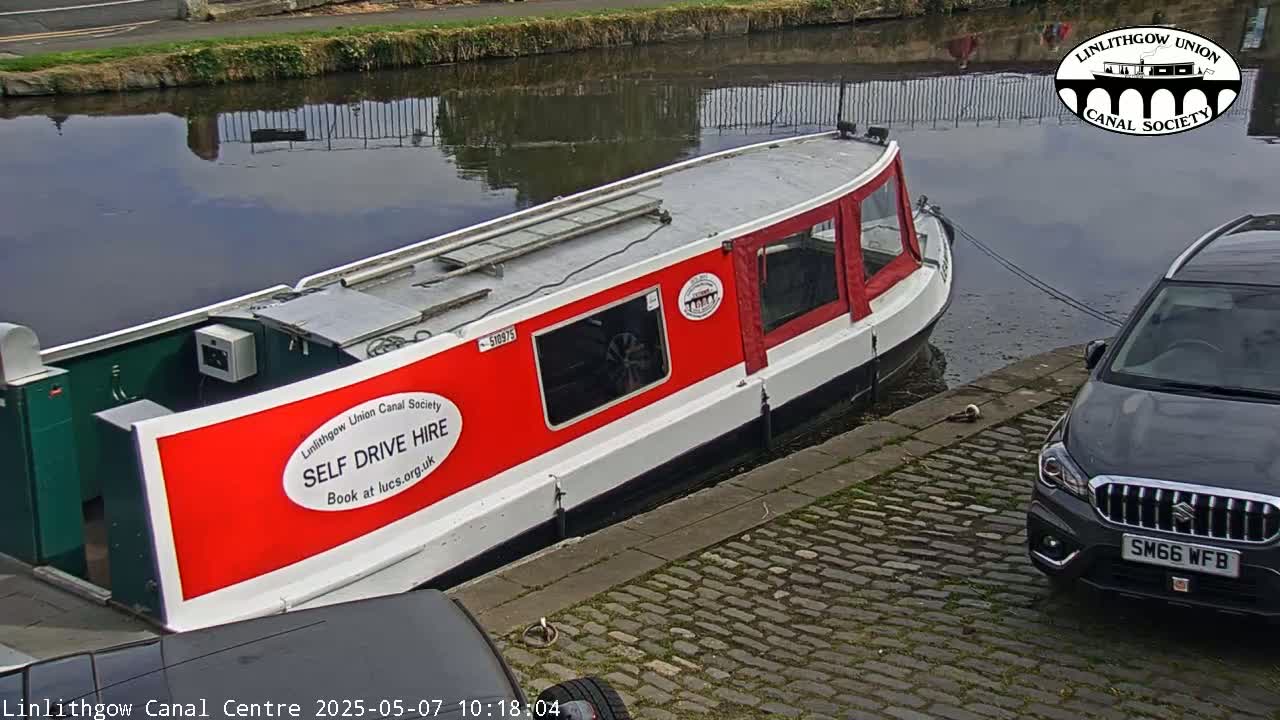 A red and white canal boat is moored alongside a cobblestone embankment next to a dark-colored SUV, with calm water reflecting the sky in the background.