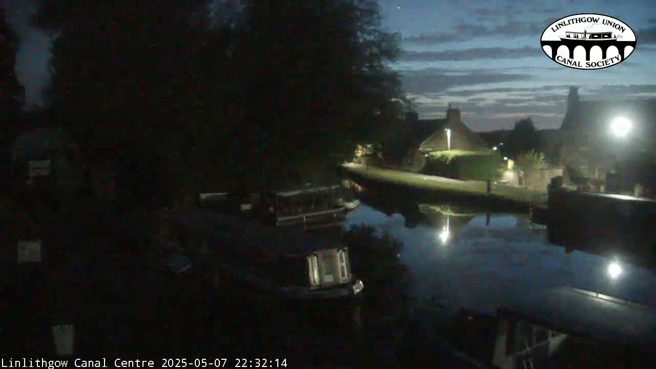 A dark canal at night, under a partly cloudy sky, reflects lights from buildings and boats moored along its banks.