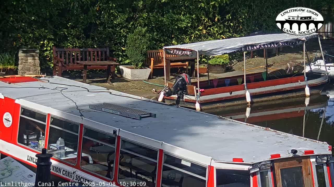 Two red canal boats are moored on a sunny day, next to a stone wall, wooden benches and some shrubbery.