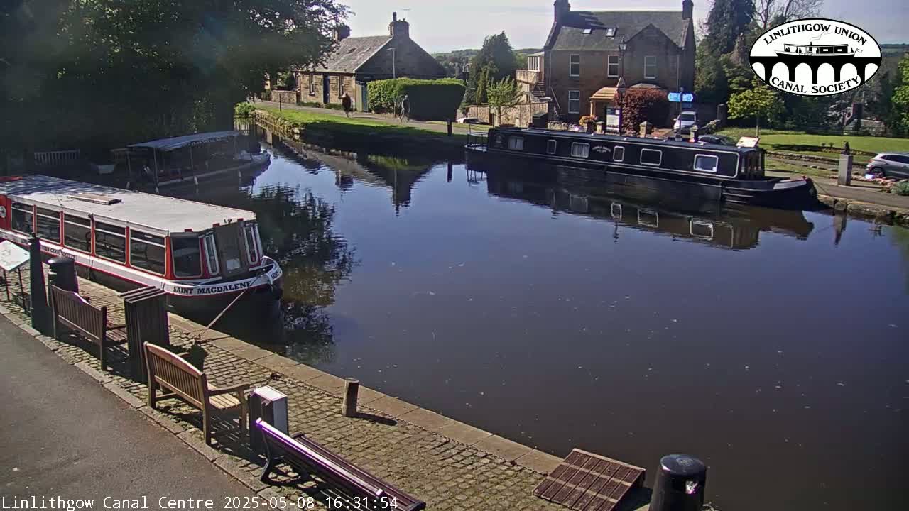 On a sunny day, several boats are moored along a calm canal next to stone buildings and green vegetation.