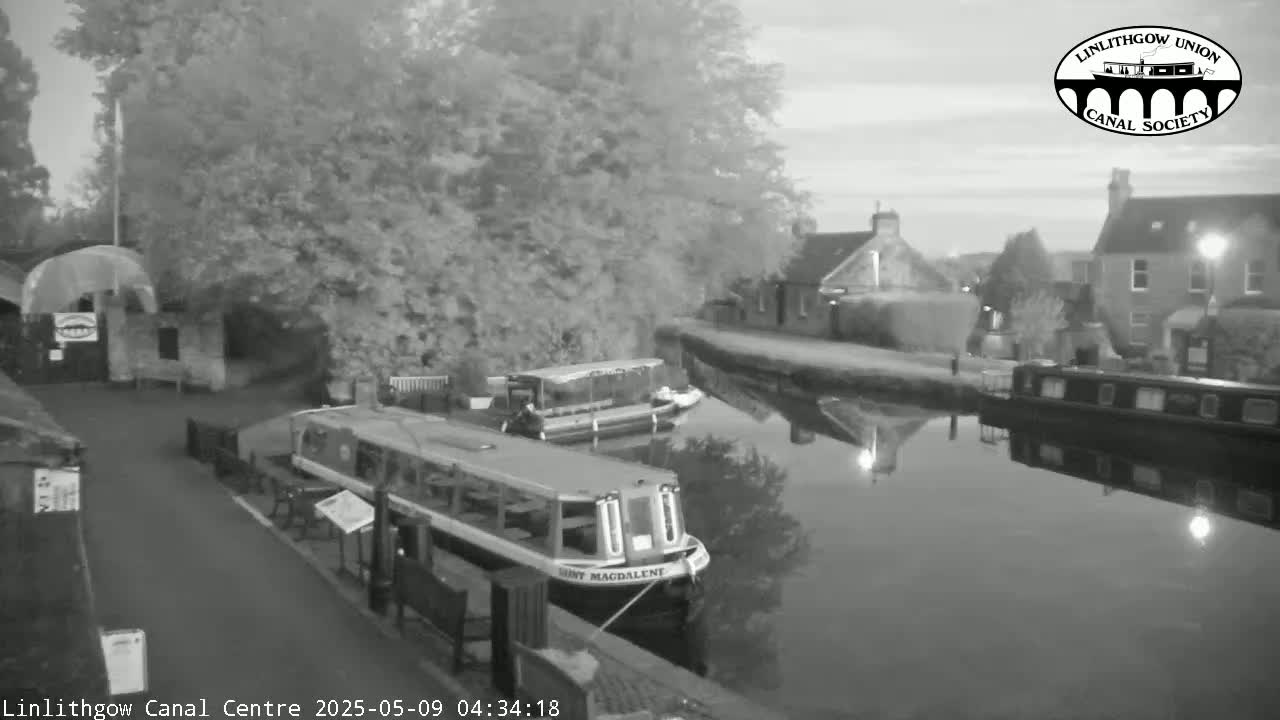 A calm canal at dawn, lined with trees and buildings, features two moored canal boats.