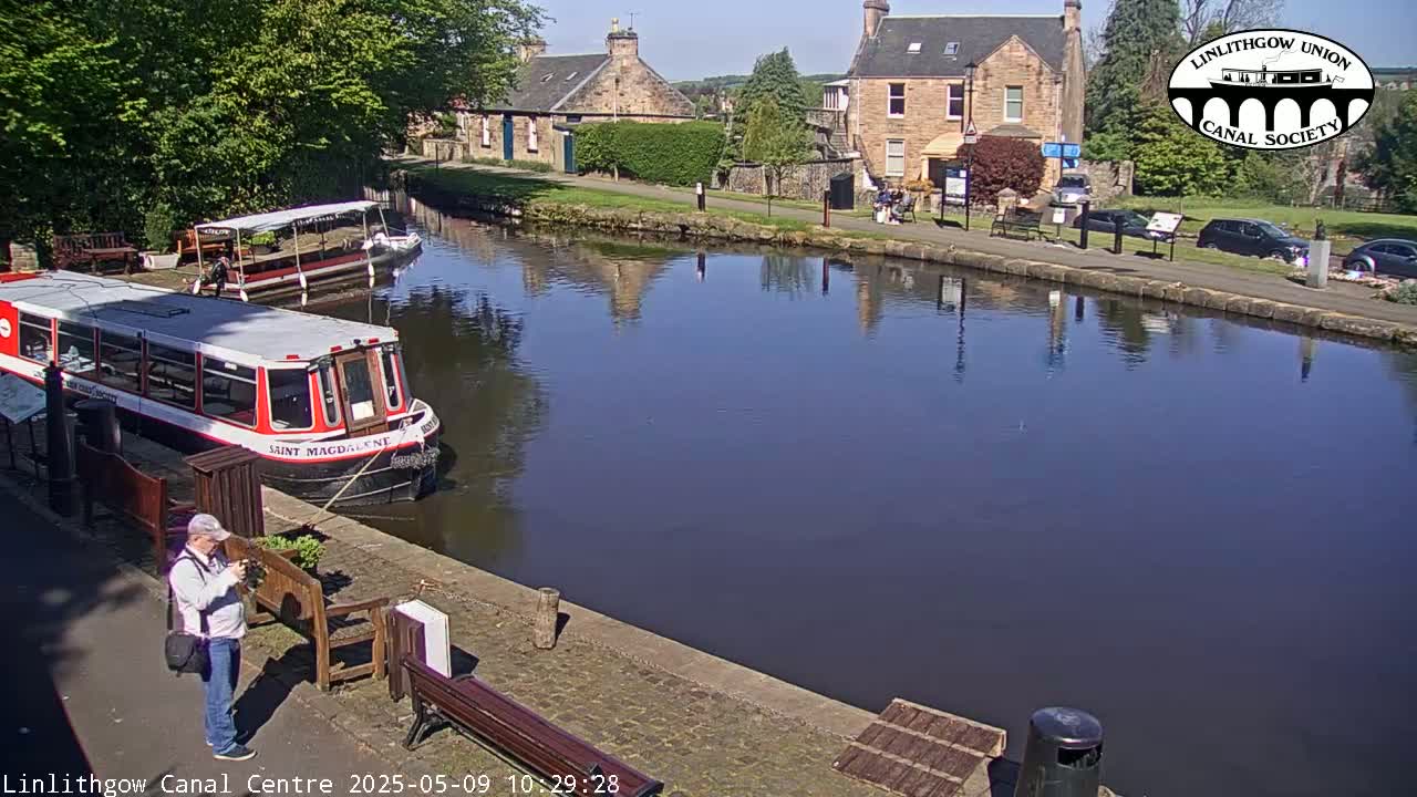 Two canal boats are moored beside a calm canal, next to stone buildings and a paved walkway on a sunny day.