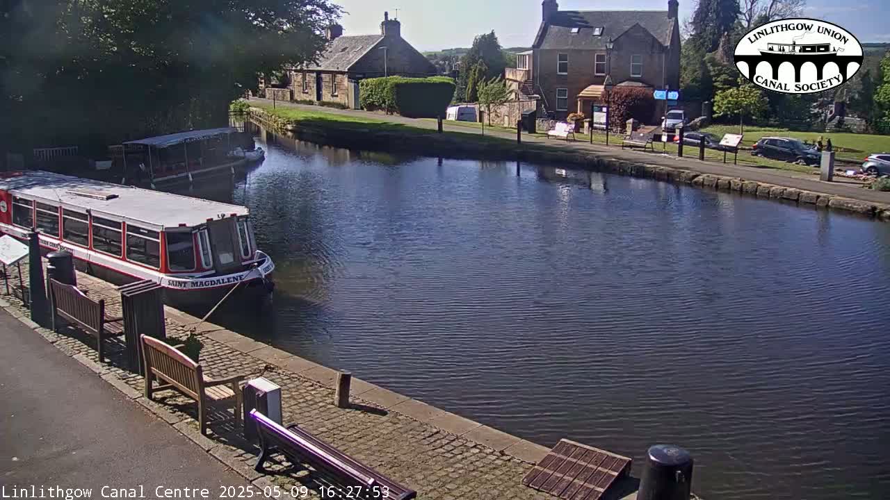 A canal scene on a sunny day shows two canal boats moored alongside a paved walkway, with stone buildings and some parked cars visible in the background.