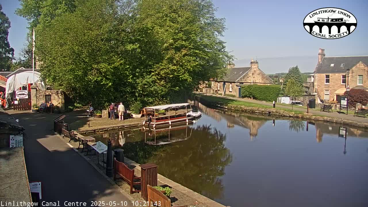 A canal reflects buildings and trees under a sunny sky, with a small boat docked and several people standing nearby.