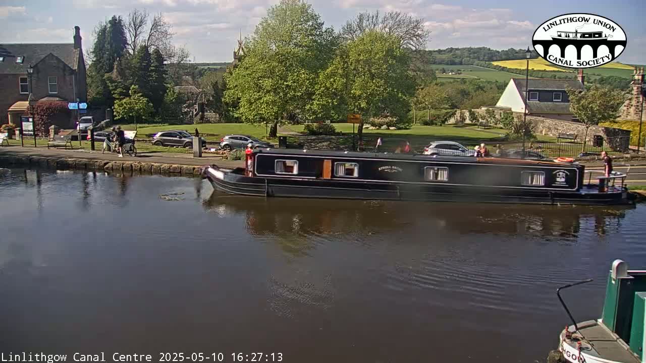 A narrowboat is moored on a canal beside a stone building and green space under a partly cloudy sky.