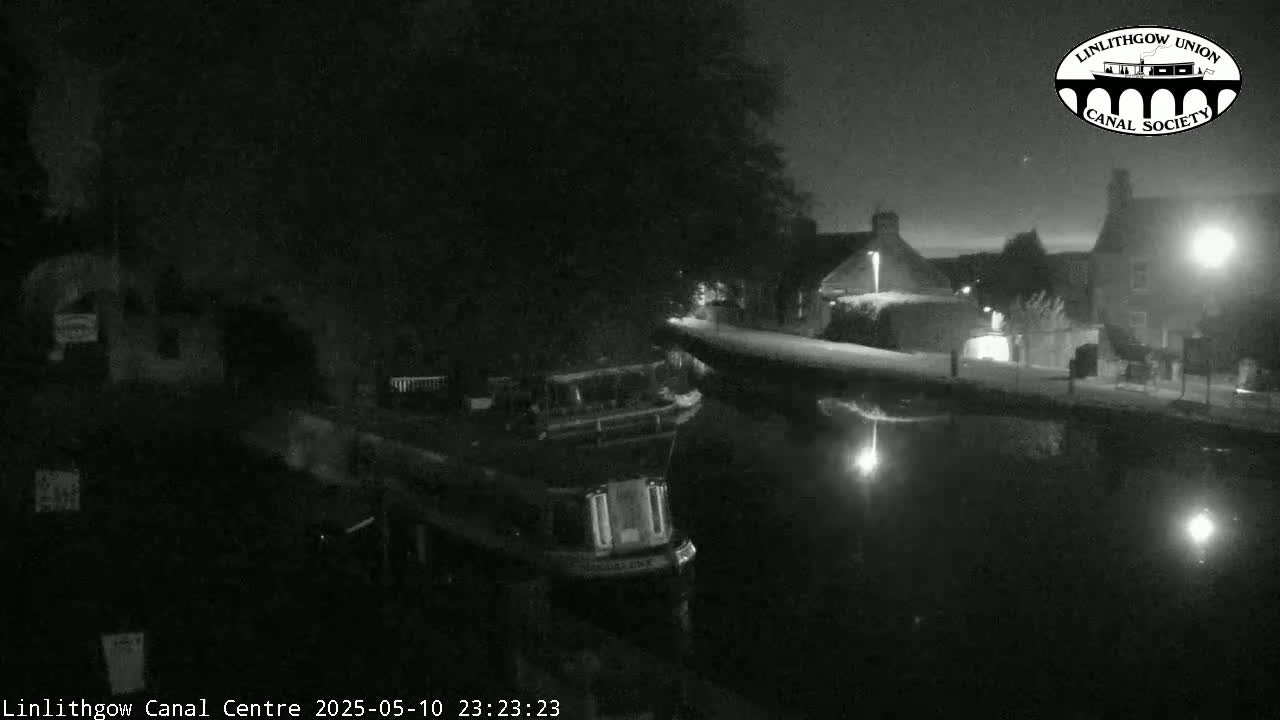 A nighttime, black and white image shows a canal with a boat moored alongside a snow-covered embankment next to dark buildings.
