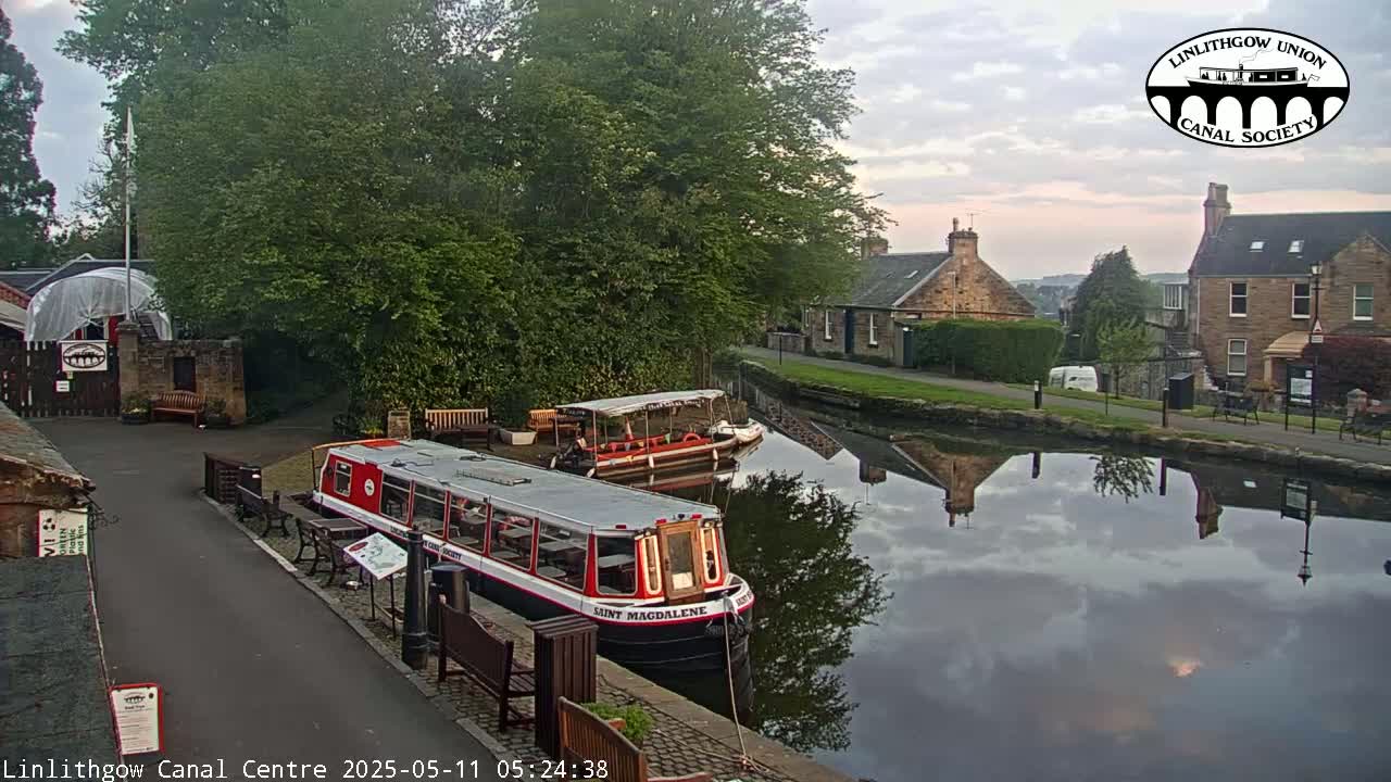 Two canal boats are moored near a canal-side path and buildings under a partly cloudy sky.