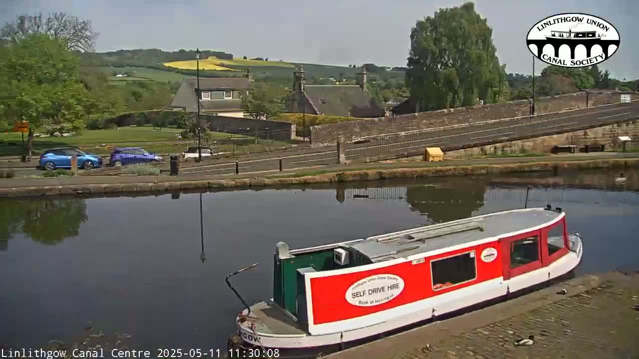 A red narrowboat is docked at a canal next to a stone bridge and buildings under a sunny sky.