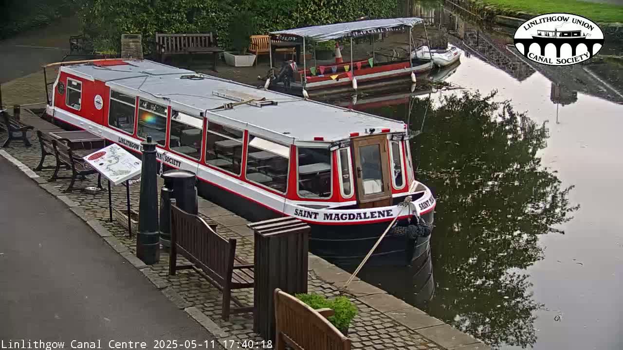 A red and white canal boat named Saint Magdalene is moored beside a dock, with another smaller boat nearby on a calm, overcast day.