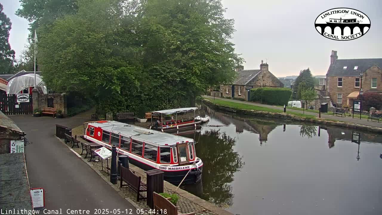 A calm canal reflects buildings and trees on an overcast day, with two canal boats docked nearby.