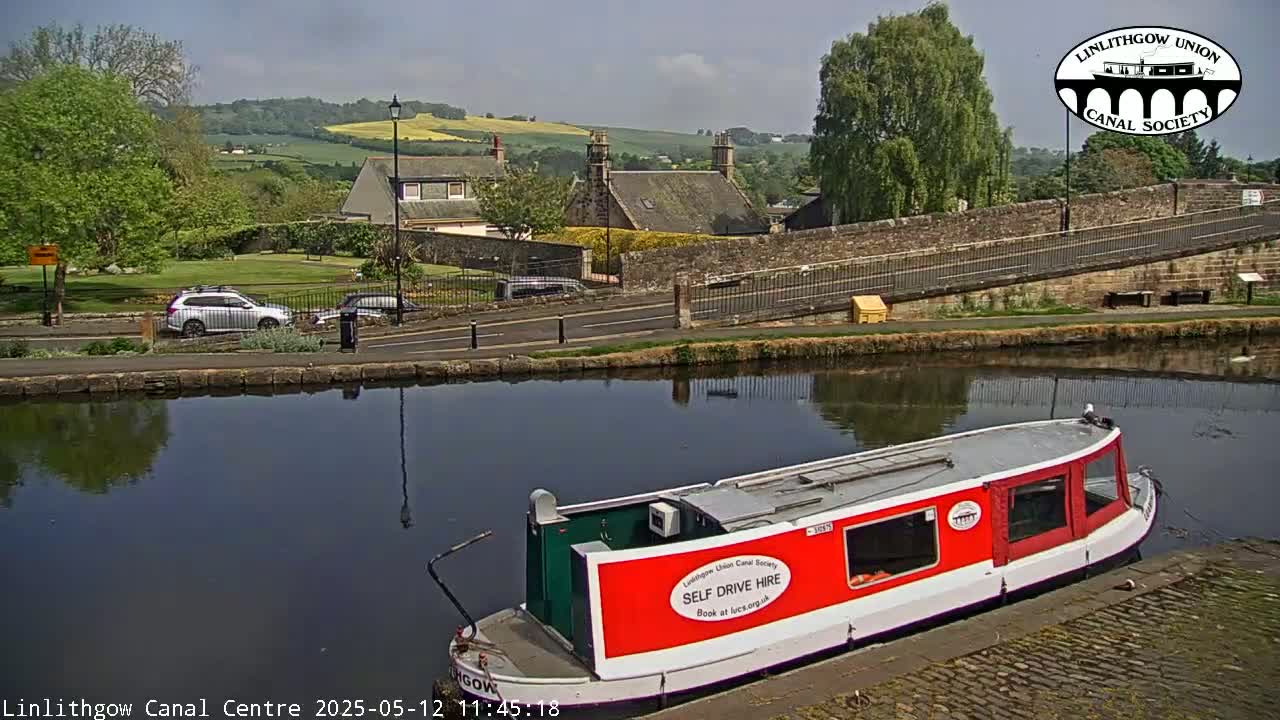 A red canal boat is moored beside a calm canal, with a stone bridge and buildings visible beyond it under a mostly sunny sky.
