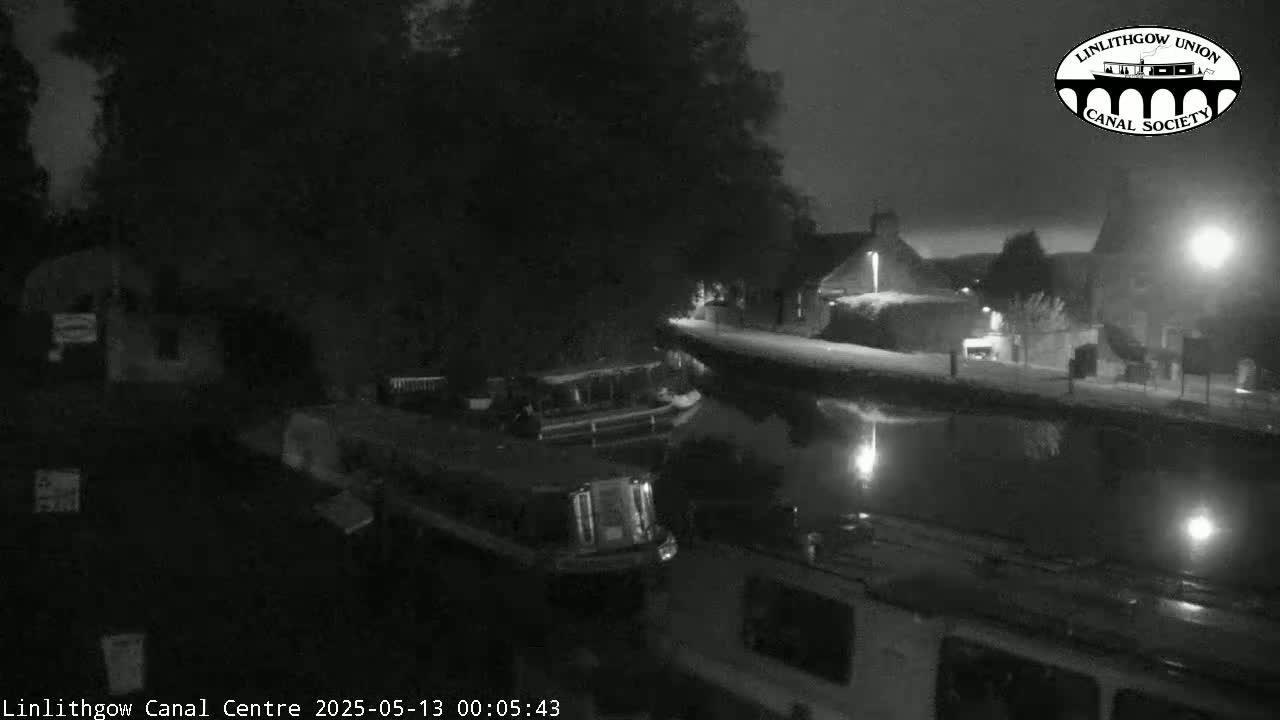 A nighttime, black-and-white view of a canal with boats moored alongside a snow-dusted embankment, under dim lighting.
