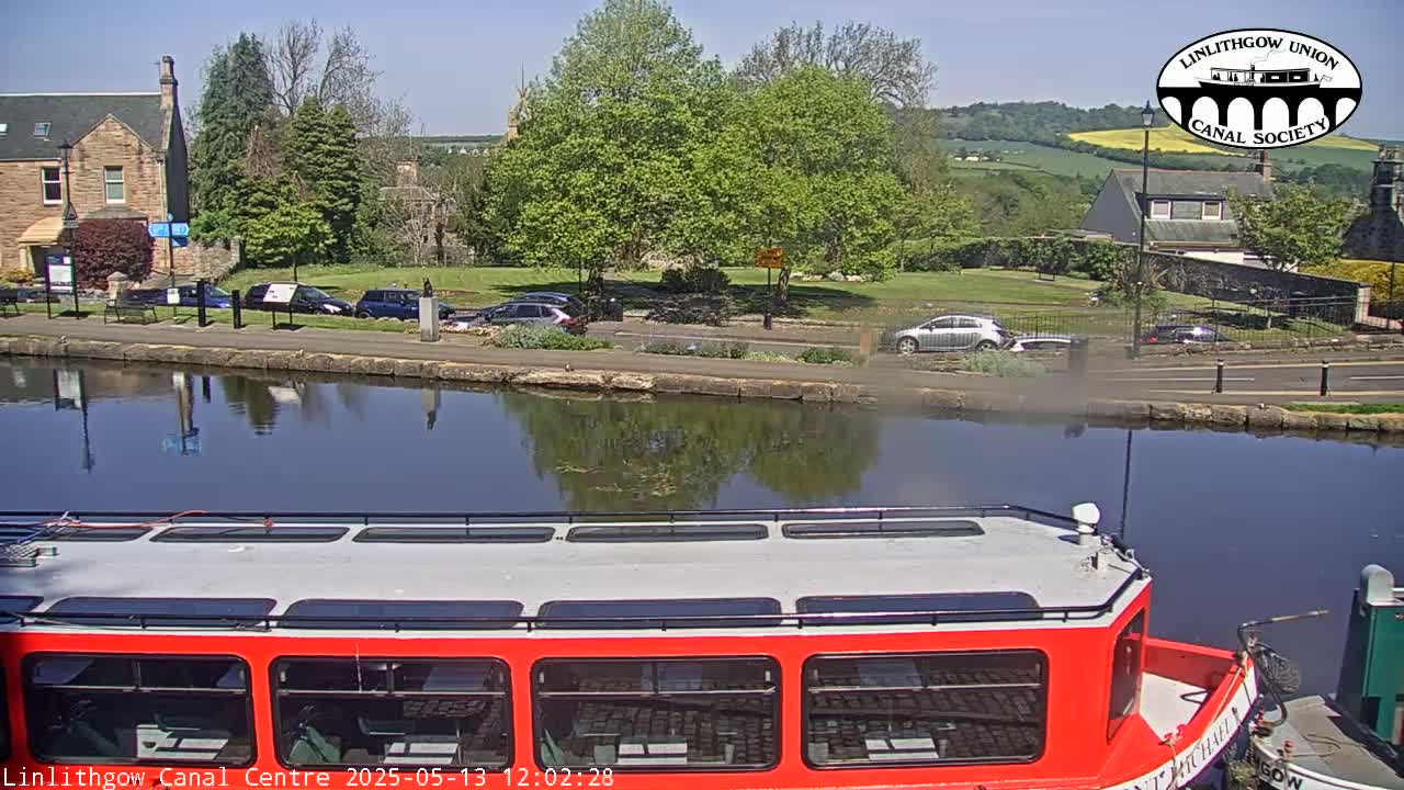 A red canal boat is moored on a calm canal under a sunny sky, with cars parked along a paved bank beside green trees and buildings in the background.
