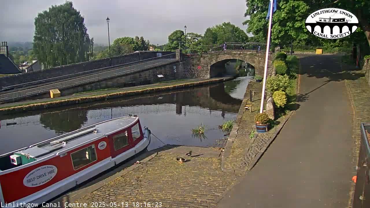 A red narrowboat is moored at a canal side, under a partly cloudy sky, near a stone arched bridge and towpath.