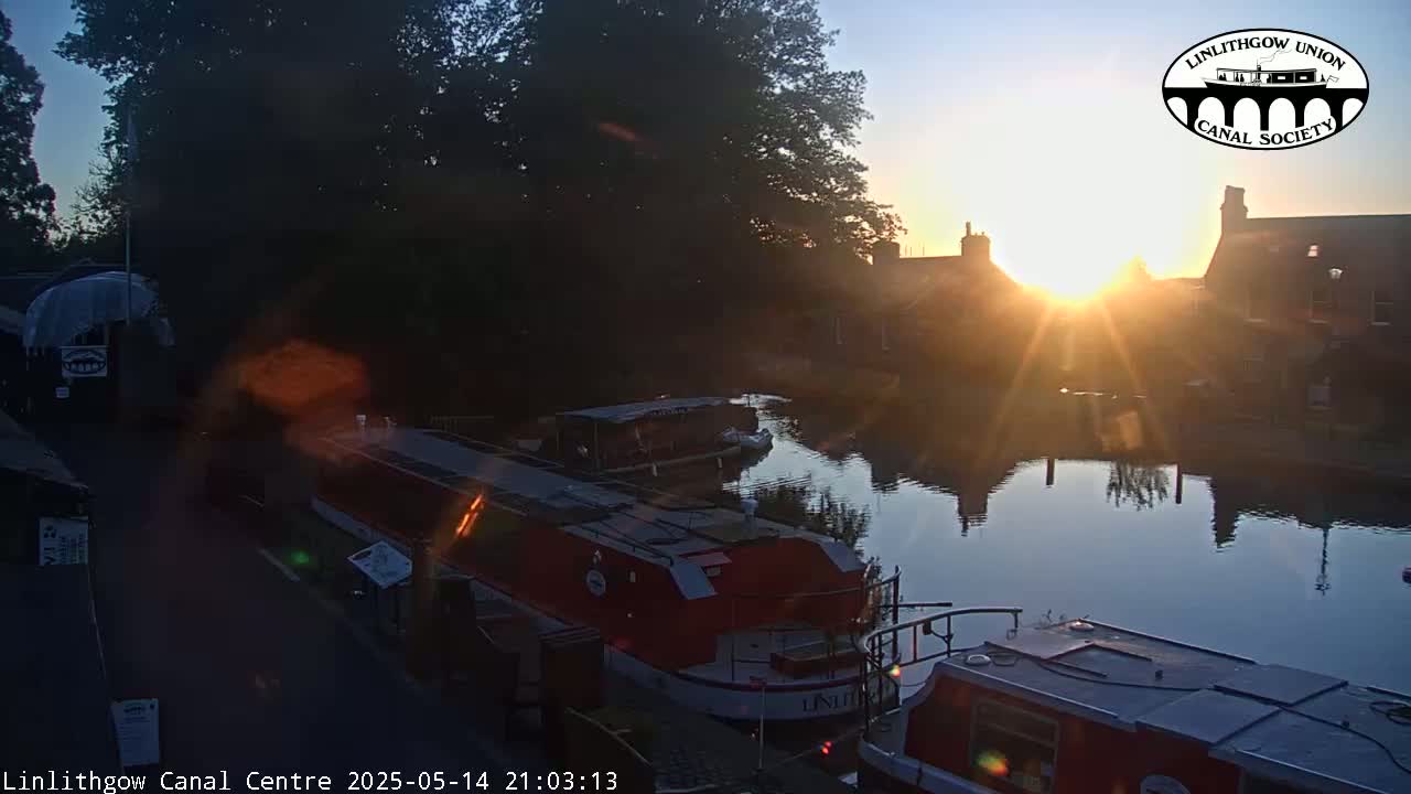 A canal at sunrise, with several narrowboats moored alongside the bank, under a mostly clear sky.
