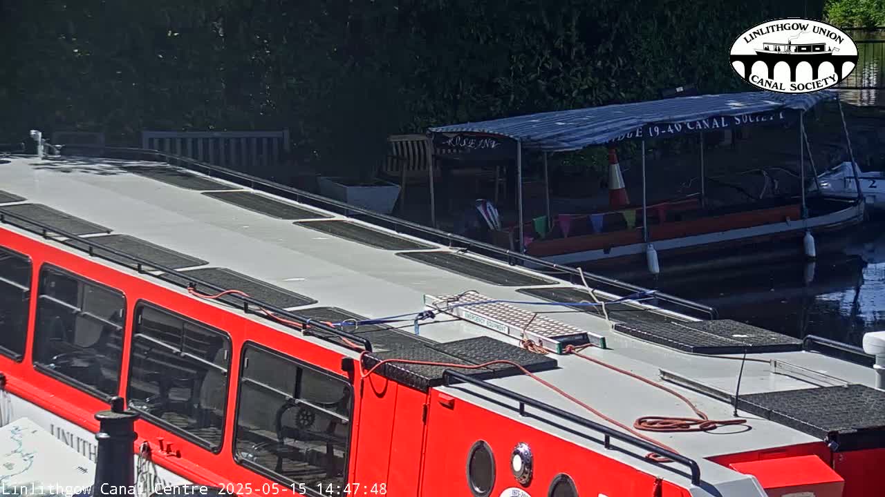 A red and gray canal boat is moored next to another smaller boat on a sunny day.