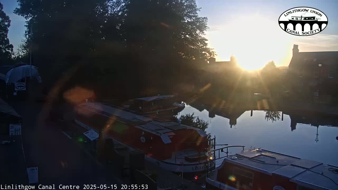 Several boats are moored on a calm canal at sunrise, with buildings and trees lining the waterway.