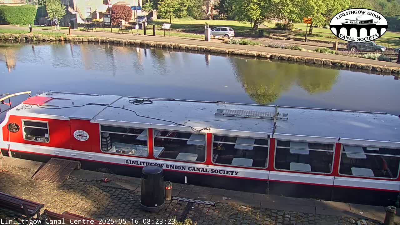 A red and white canal boat is docked beside a calm canal reflecting the surrounding trees and buildings under a sunny sky.