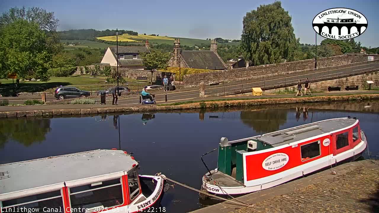 Two canal boats are moored next to a stone bridge on a sunny day, with a calm canal reflecting the surrounding buildings and greenery.