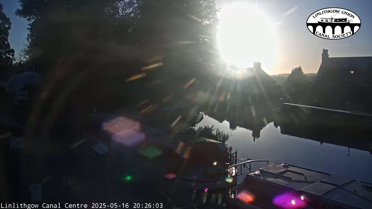 Bright sunlight shines over a calm canal, reflecting off the water and illuminating boats moored along the bank, with buildings and trees visible in the background.