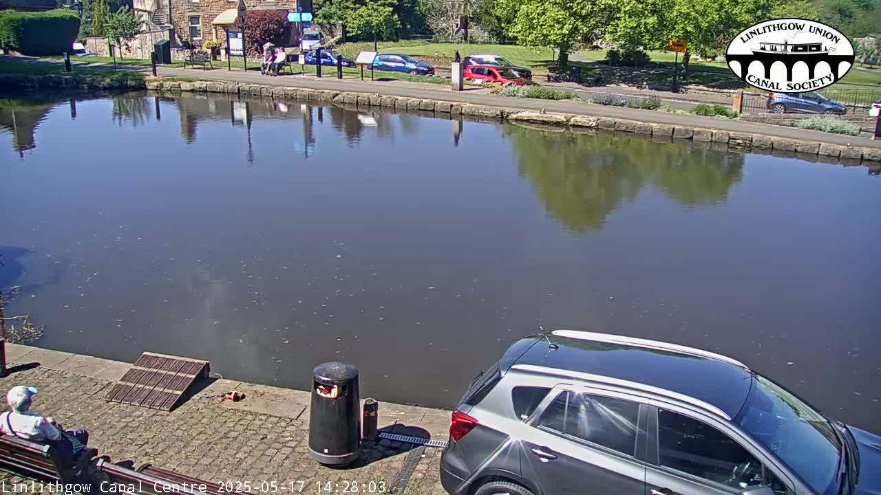 A calm canal reflects buildings and trees under a sunny sky, with a silver SUV parked by the water's edge and a person sitting on a bench.