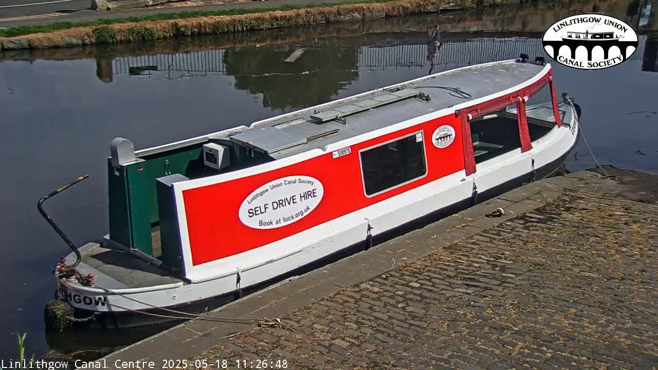 A red and white narrowboat is moored alongside a cobblestone path next to a calm canal on a sunny day.