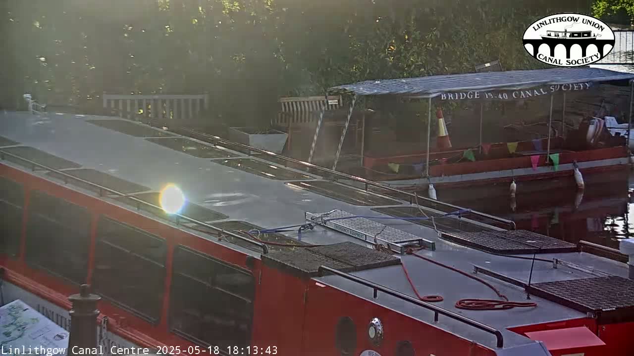 The top of a red canal boat is visible, along with another boat behind it, on a calm canal under a hazy, sunny sky.