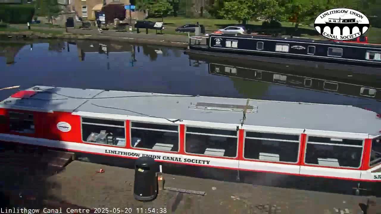 A red and white canal boat is docked next to a black canal boat on a calm canal, under a sunny sky.