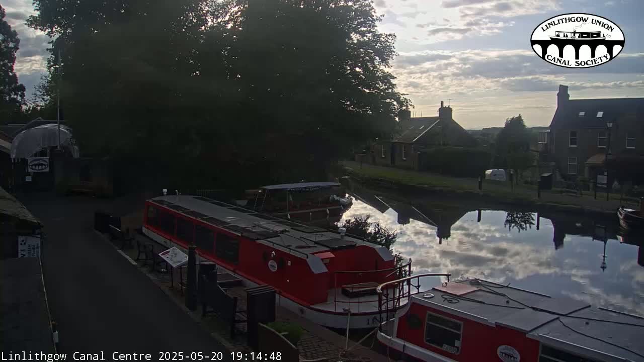 A calm canal reflects a partly cloudy sky at sunset, with several red narrowboats moored alongside a paved walkway and buildings in the background.