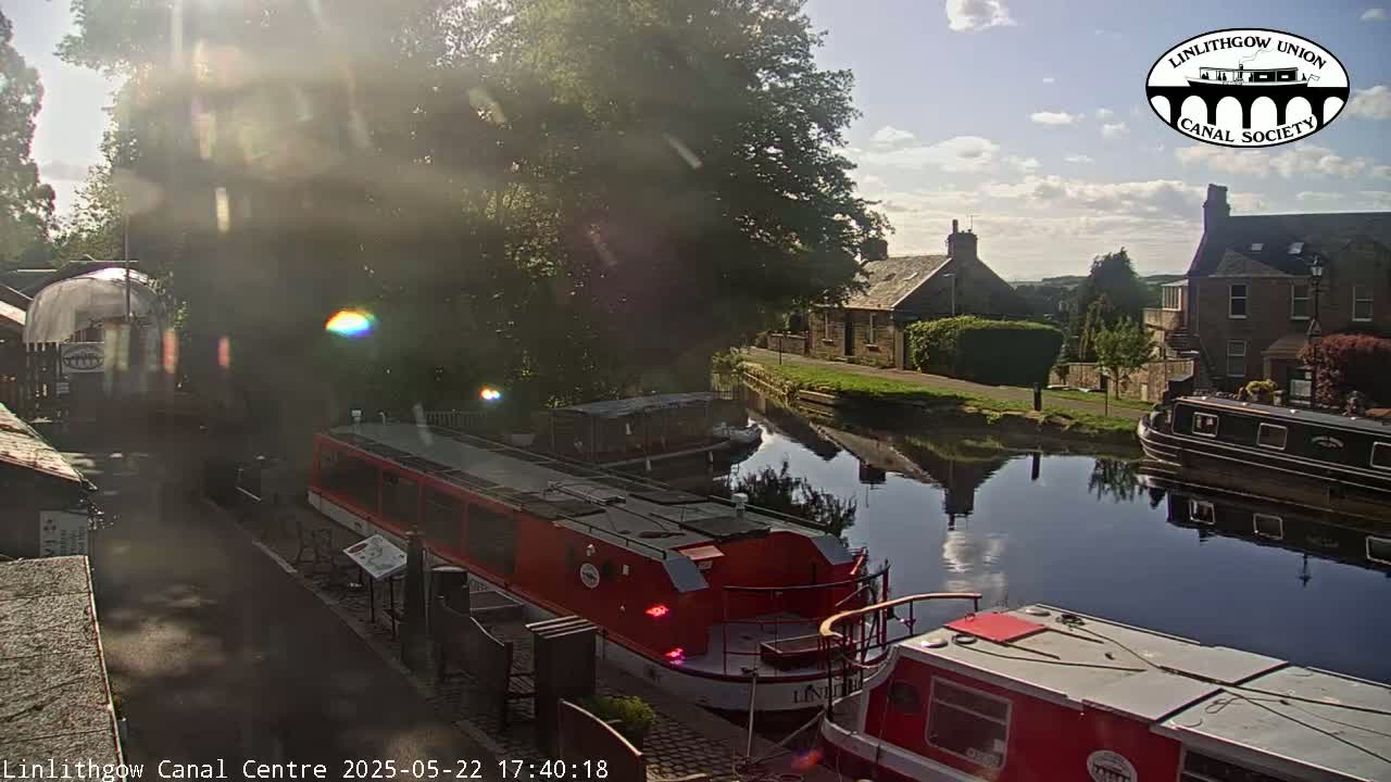 Sunlight streams through trees onto a canal where several narrowboats are moored, some buildings are visible in the background on a sunny day.