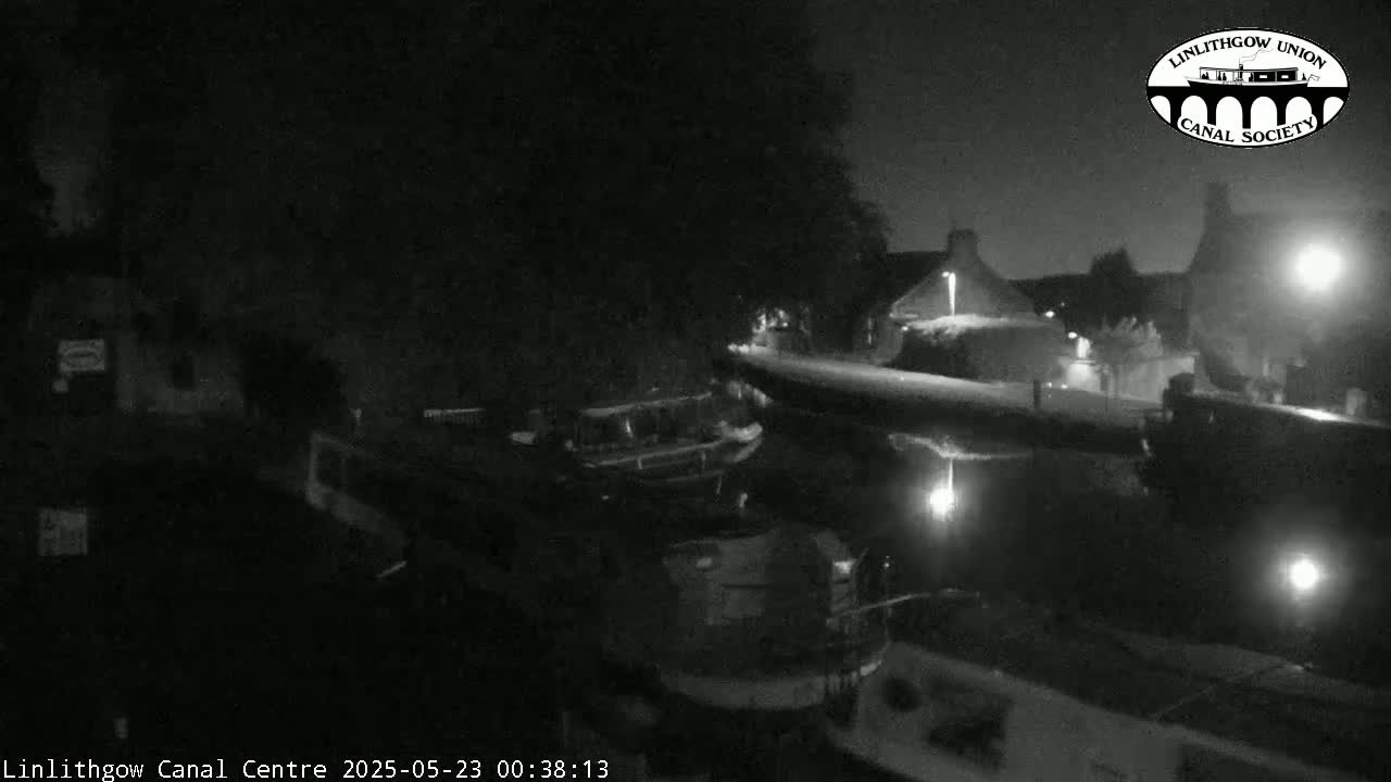 A nighttime, black-and-white view of a canal scene with boats moored alongside buildings under a dark sky.