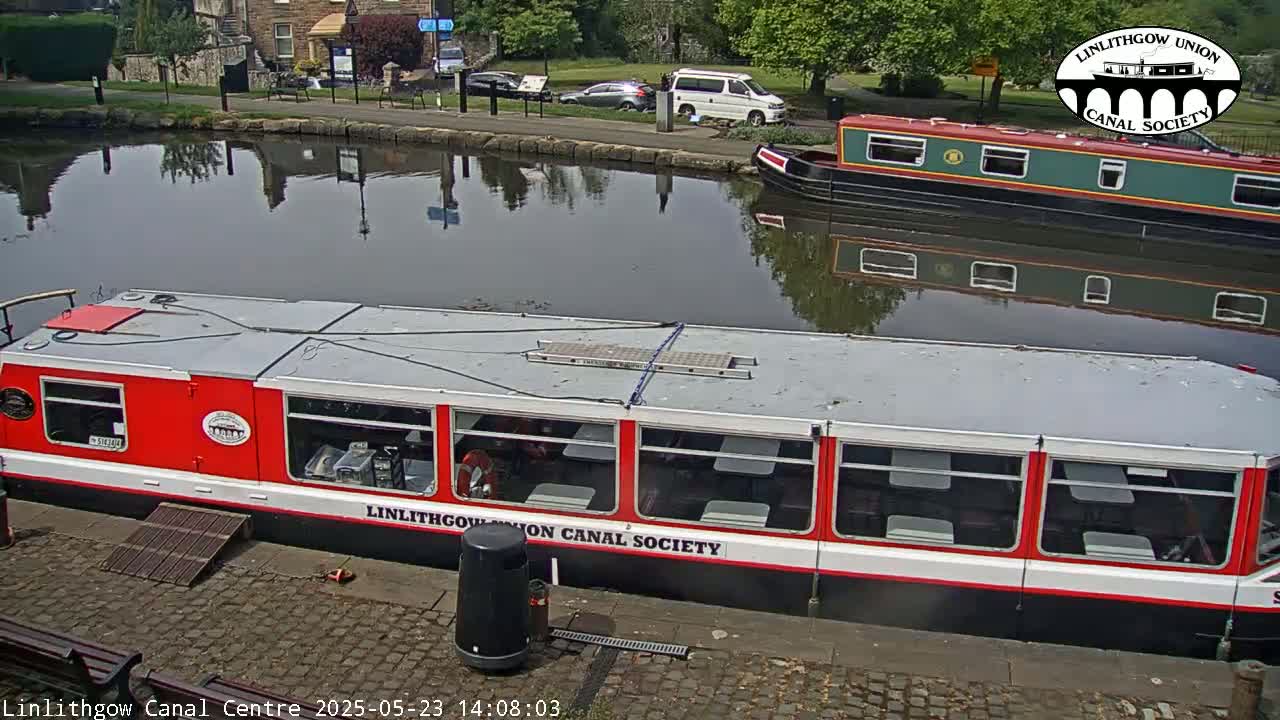 A red and white canal boat is docked next to a smaller, green and red canal boat, on a sunny day.