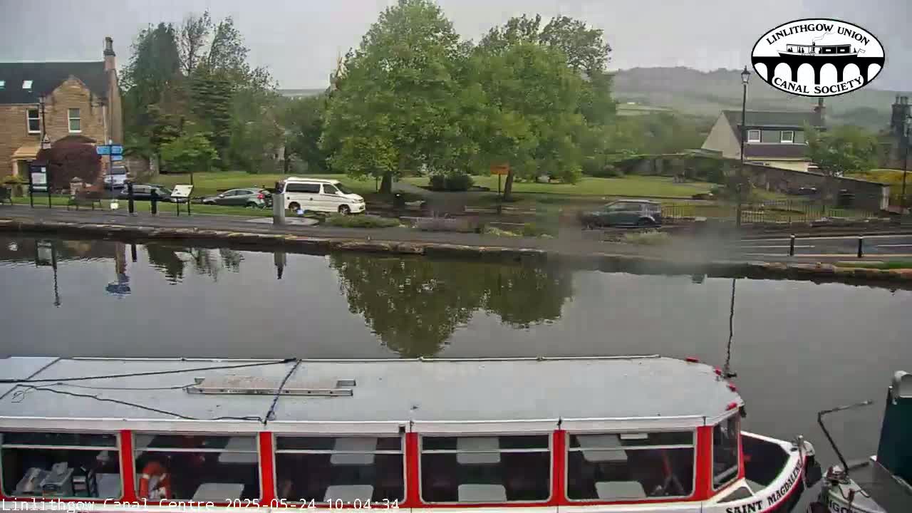 A canal scene on an overcast day shows a red and white passenger boat in the foreground, with several vehicles parked along the canal bank and buildings in the background.