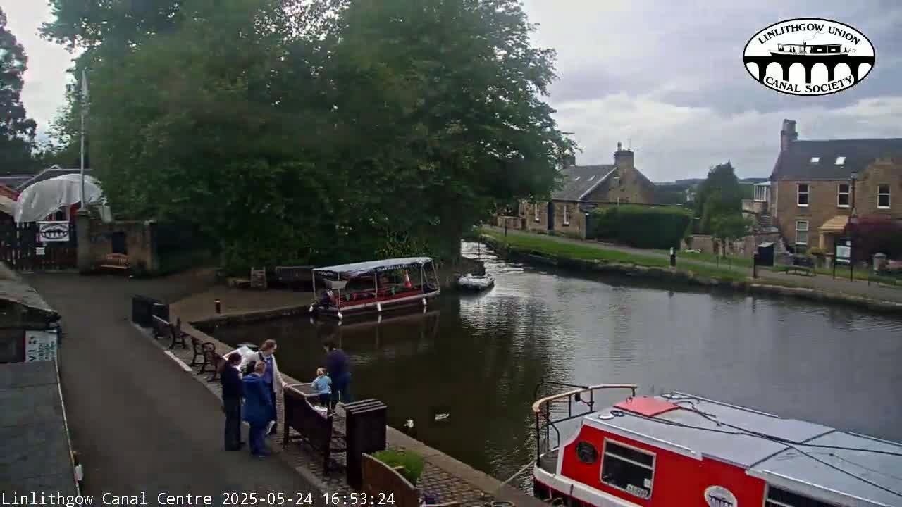 A canal scene on an overcast day shows two boats moored near a stone building, with several people standing on a paved walkway nearby.