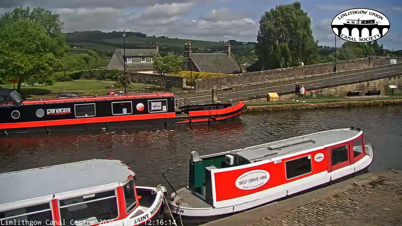 Three red canal boats are moored on a waterway under a partly cloudy sky, with a stone bridge and buildings visible in the background.