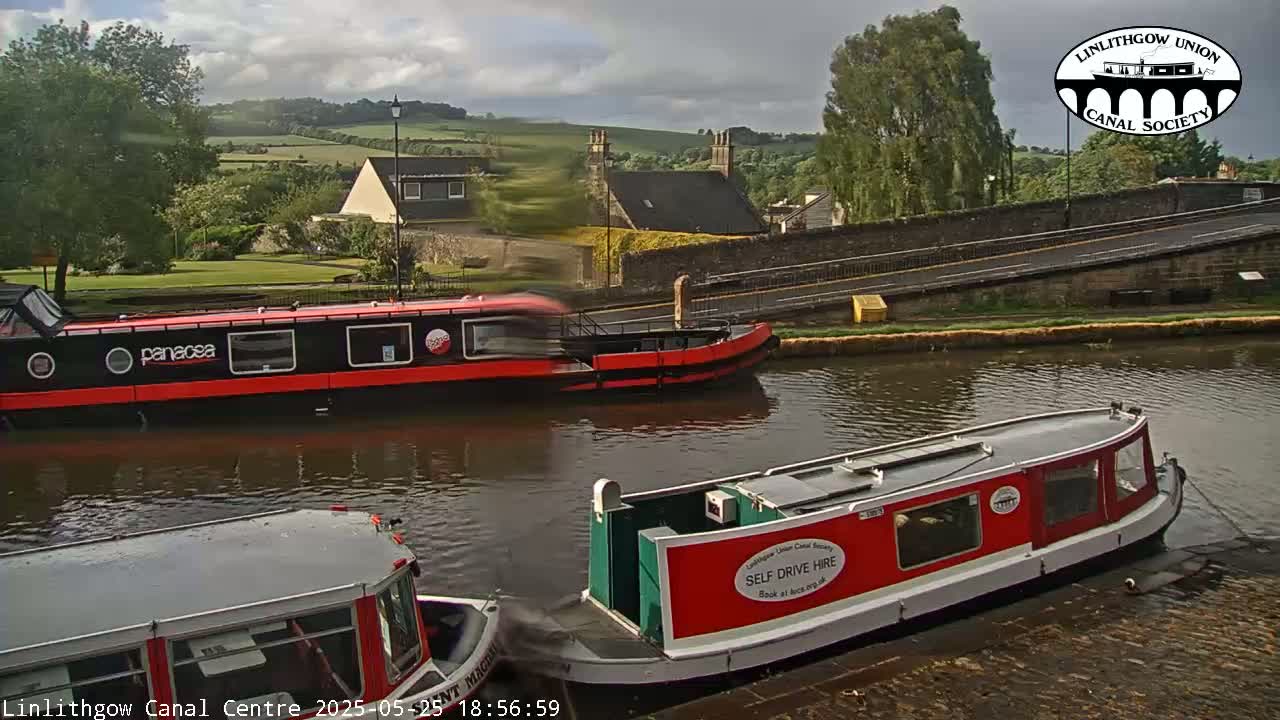 Three narrowboats are moored on a canal under a partly cloudy sky, with green hills and buildings visible in the background.
