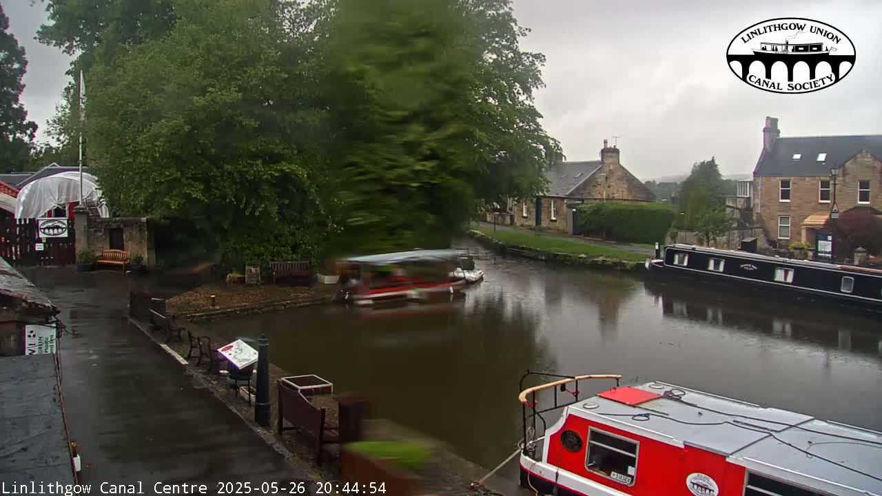 A canal scene on a rainy day shows boats moored along a waterway, near buildings and lush greenery.