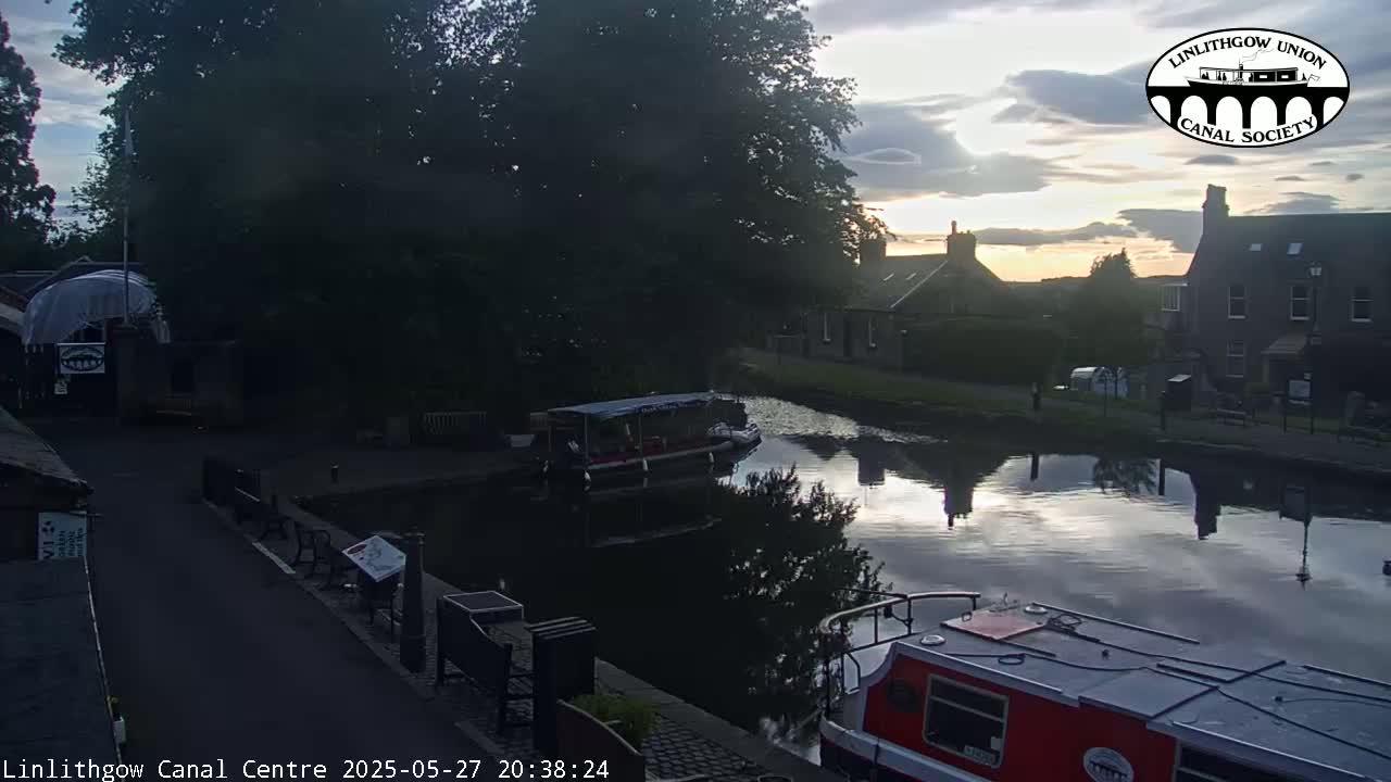 A calm canal reflects a partly cloudy sunset sky, with boats moored along its banks near buildings.