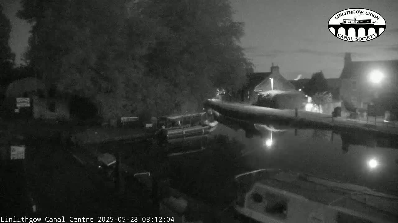 A nighttime, black-and-white view of a canal, with boats moored along its banks, under a dark sky.