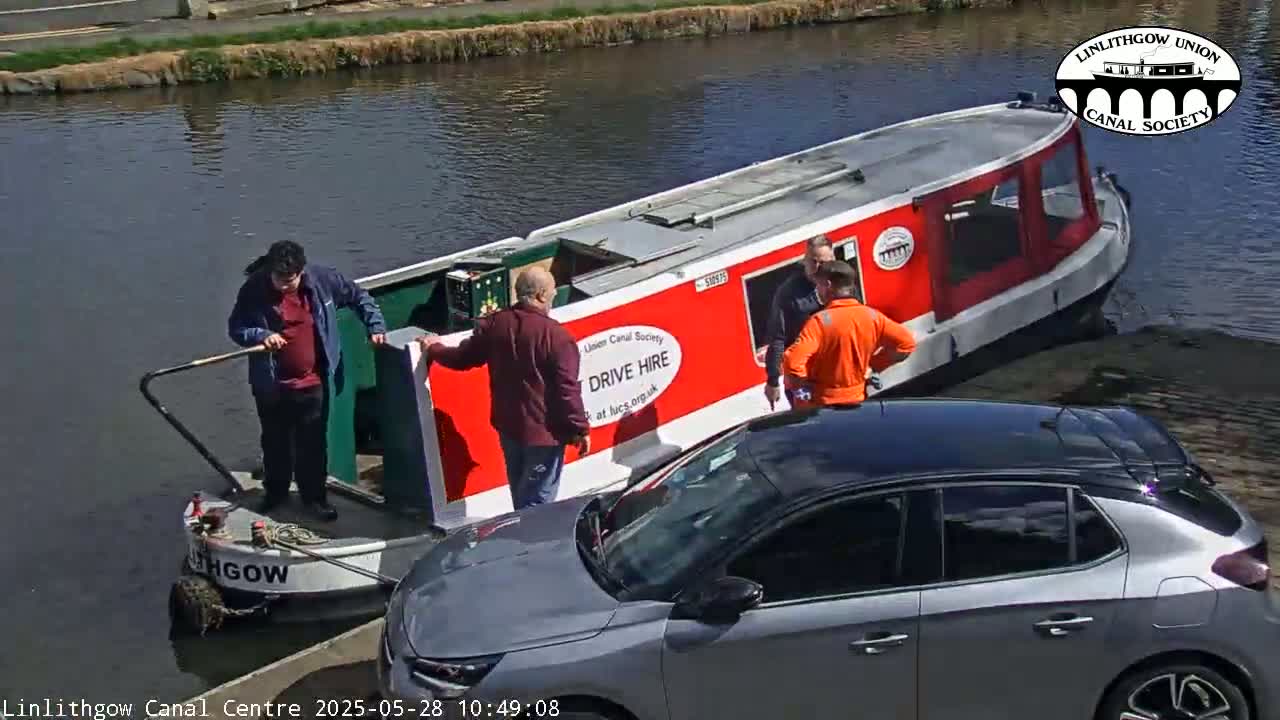 Three people are loading a large sign onto a red canal boat, next to a gray car, on a sunny day.