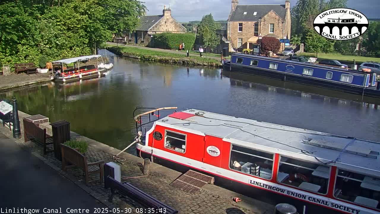 A sunny day shows several boats on a canal, with buildings and trees lining the waterway.