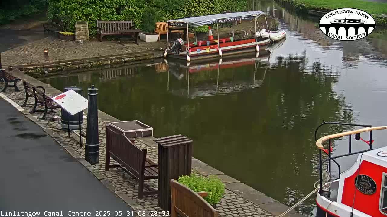 A calm canal reflects the overcast sky, with several benches lining the paved bank, and two boats, one a small passenger vessel and the other a narrowboat, moored nearby.