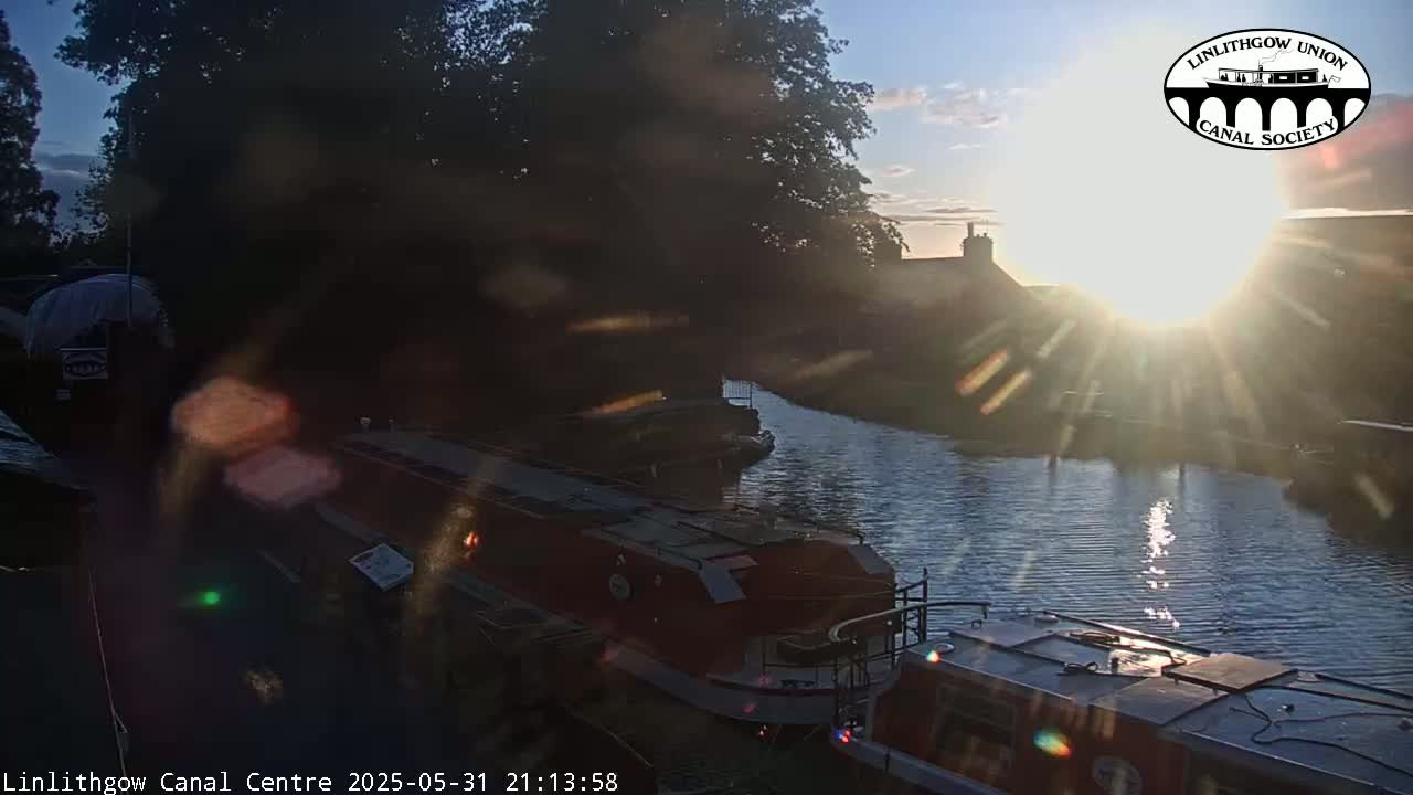 A bright sunset shines over a canal, where several boats are moored alongside the bank, under the shade of trees.