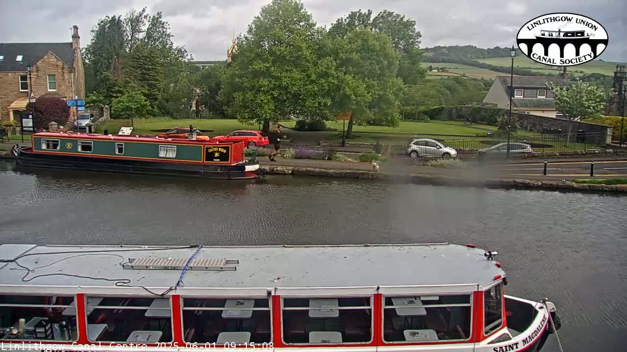 A canal scene on an overcast day shows two narrowboats, one docked and one approaching, with cars parked along a roadway next to a grassy area and buildings in the background.