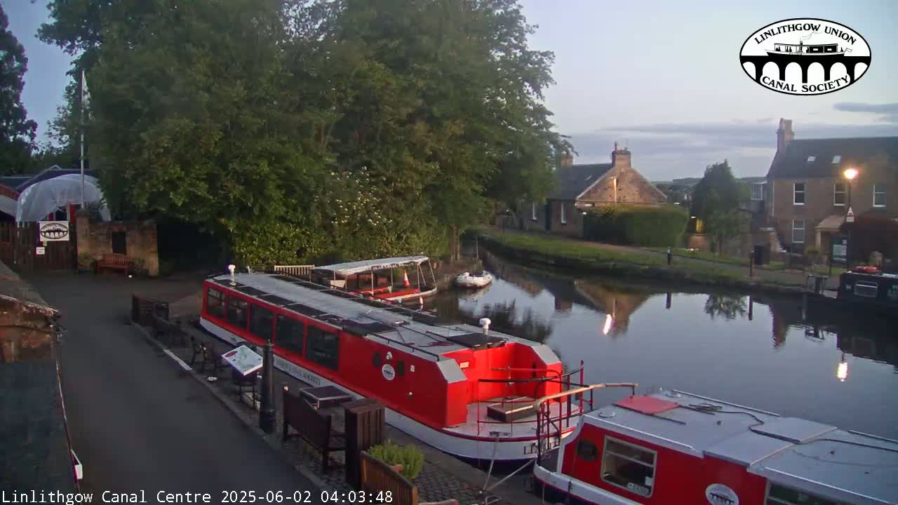 Two red canal boats are moored alongside a canal, under a twilight sky, near buildings and lush green trees.